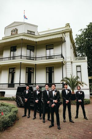 Wedding party in formal black attire posed in front of a white colonial-style mansion.