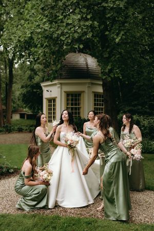 Bridesmaids in sage green dresses gathered on garden grounds with floral bouquets.