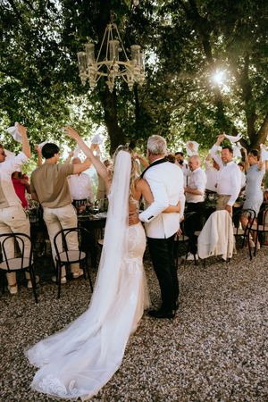 Wedding celebration scene with guests in white attire dancing under tree canopy.