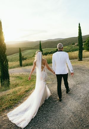 Couple in wedding attire walking away on a gravel path with cypress trees and rolling hills.