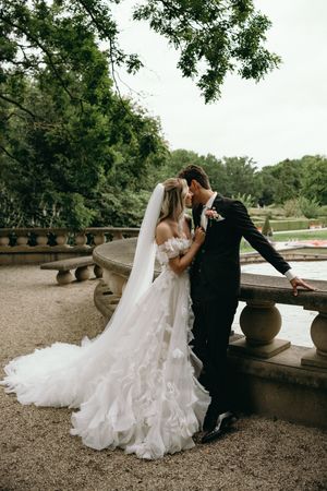 Romantic wedding couple in formal attire posing on stone balcony surrounded by trees.