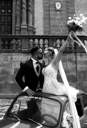 Artistic black and white photo of newlyweds posing with vintage car outside historic building.
