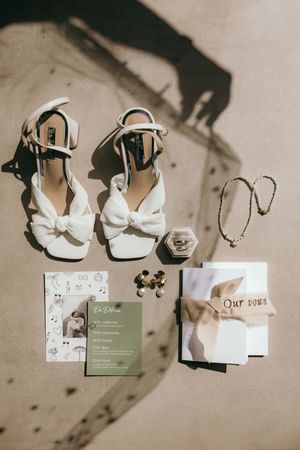 Wedding invitation suite with white shoes and jewelry arranged on a neutral background.