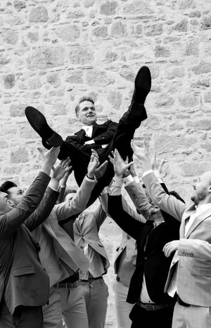 Wedding party throwing hands up in celebration against stone wall backdrop.