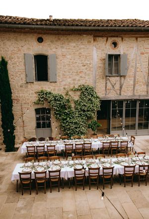 Long rustic dining table setup with wooden chairs and white linens at an outdoor stone patio venue.