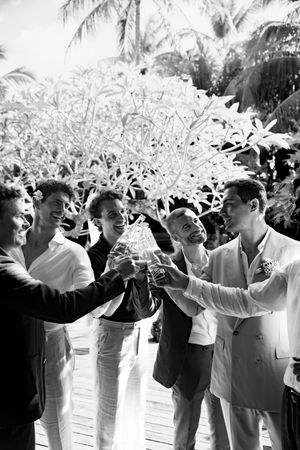 Wedding guests in light-colored attire celebrating with raised arms under palm trees.