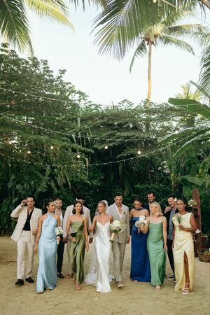 Wedding party in pastel dresses and light suits posing under palm trees on a beach.