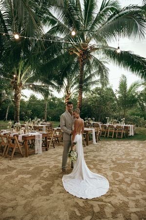 Romantic beach wedding reception setup with palm trees and wooden chairs on sandy ground at sunset.