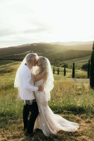 Wedding couple embracing in golden sunlit field with rolling hills and cypress trees in background.