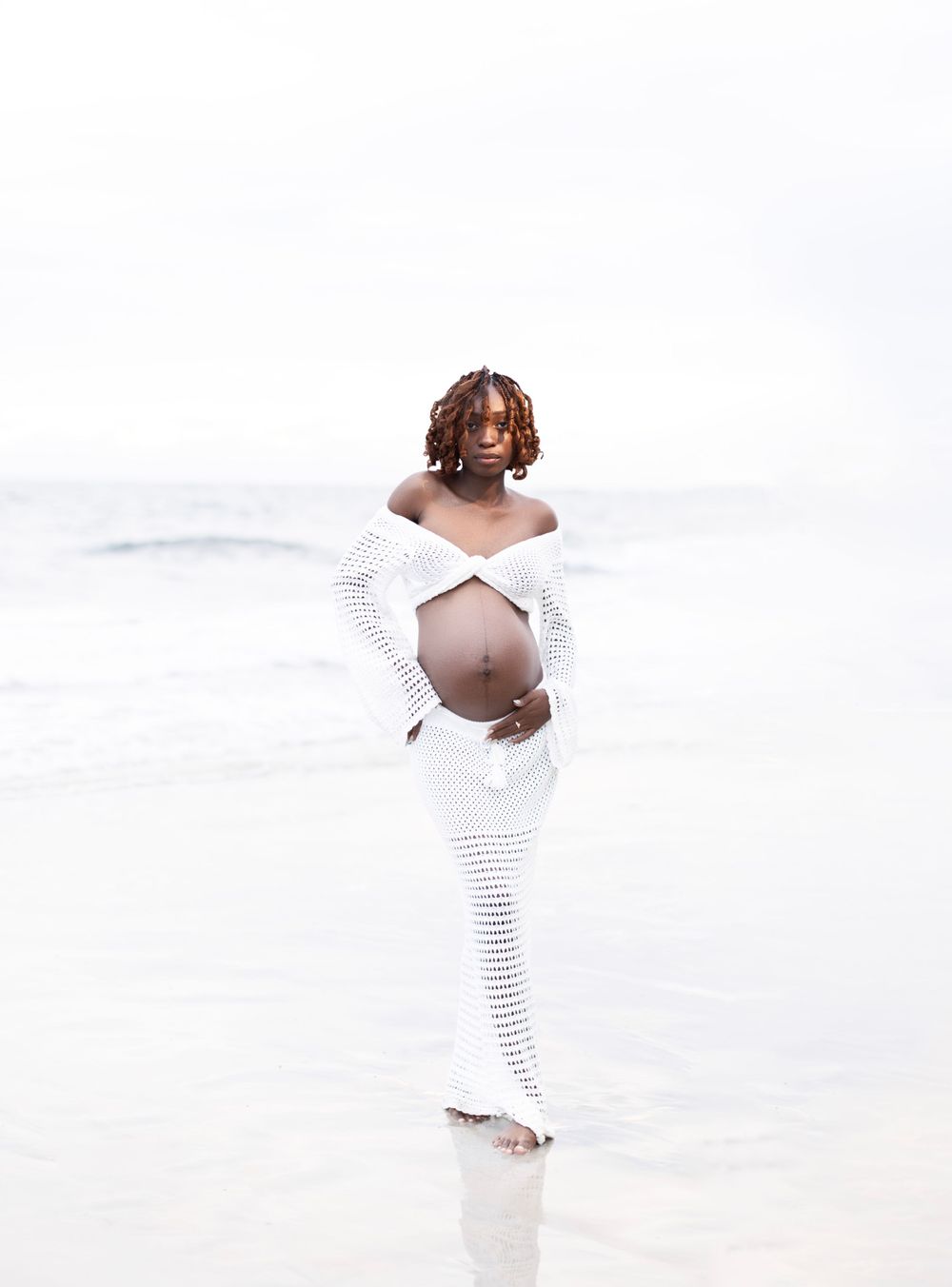 Maternity photo of expectant mother in white crochet dress on beach.