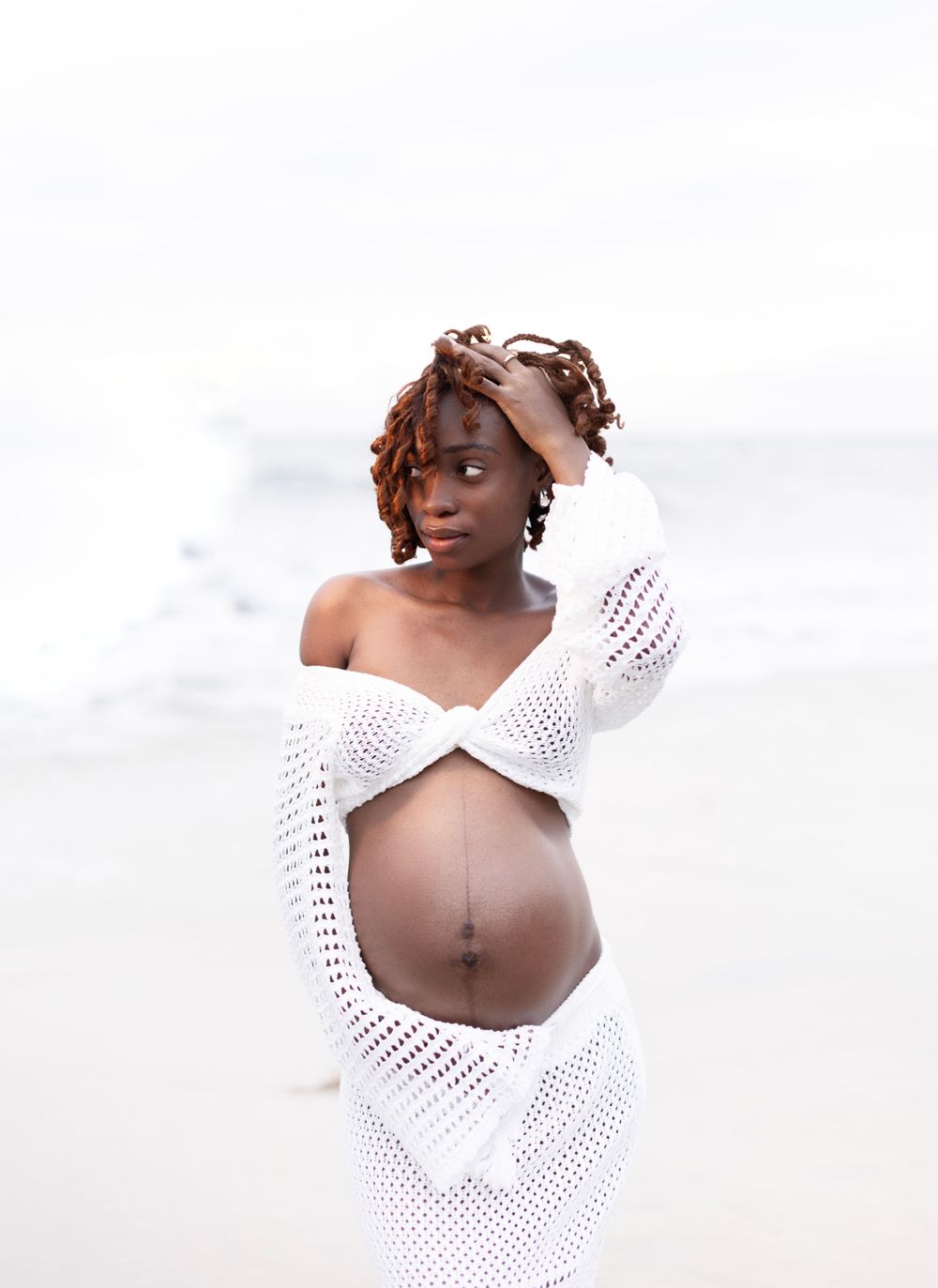 Artistic maternity portrait with woman in white lace dress against bright background.