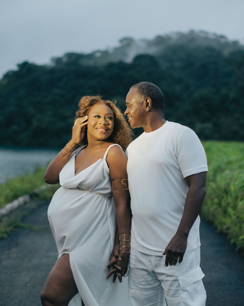 Romantic maternity photo of couple in white attire against lush mountain backdrop.