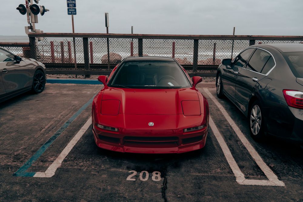 Red Honda NSX parked in spot 208 of a coastal parking lot on an overcast day.