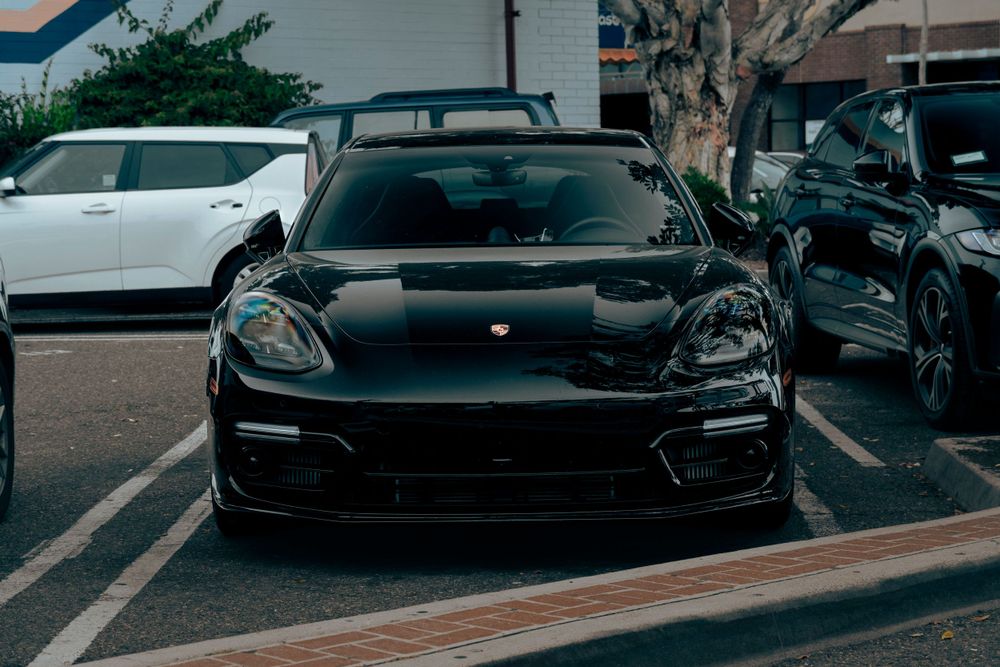 Black Porsche Panamera parked head-on in a shopping center lot between white and dark SUVs.