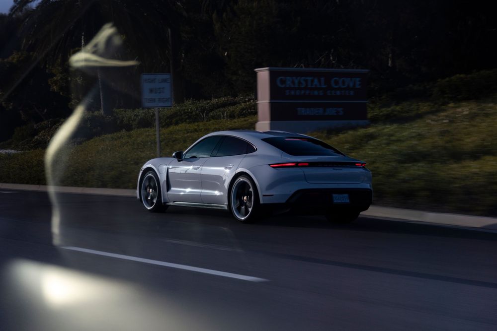 Silver Porsche Taycan Cross Turismo driving past a Crystal Cove sign on a scenic coastal road.
