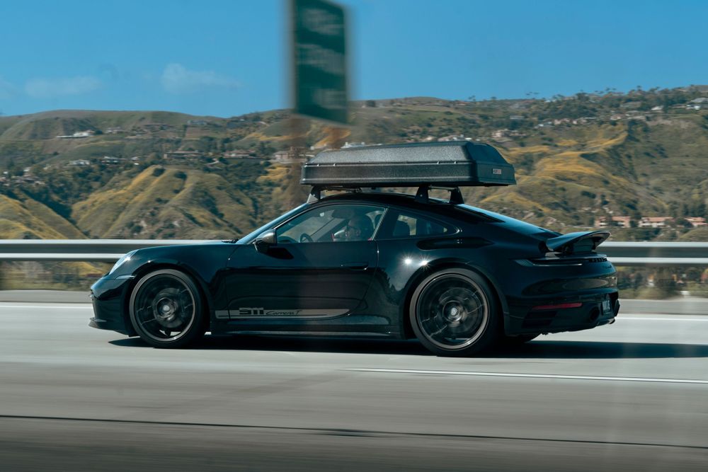Dark green Porsche 911 with roof cargo box driving on a highway with hills in the background.