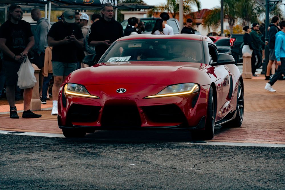 Red Toyota GR Supra at an outdoor car meet surrounded by spectators at dusk.