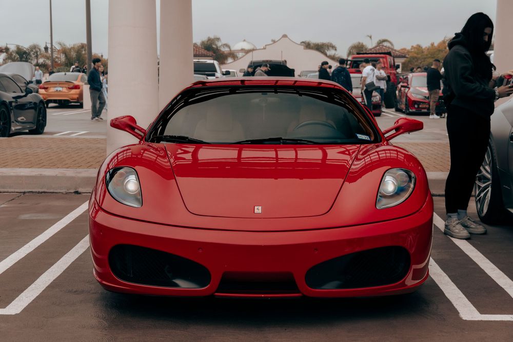 Red Ferrari F430 coupe shot head-on in a parking lot surrounded by spectators at a car show.