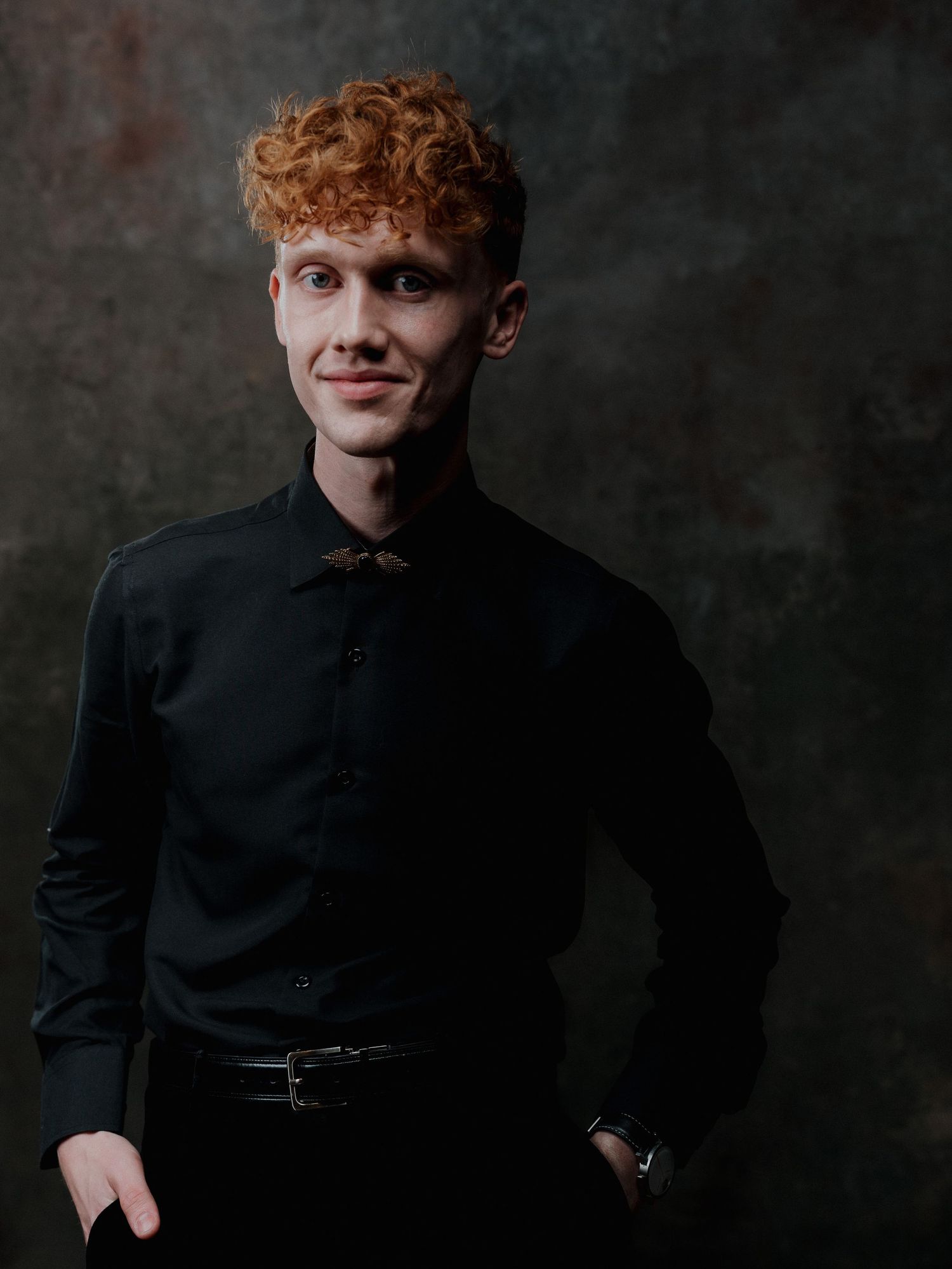 Young man with curly red hair wearing a black shirt and bow tie posing against a dark background.