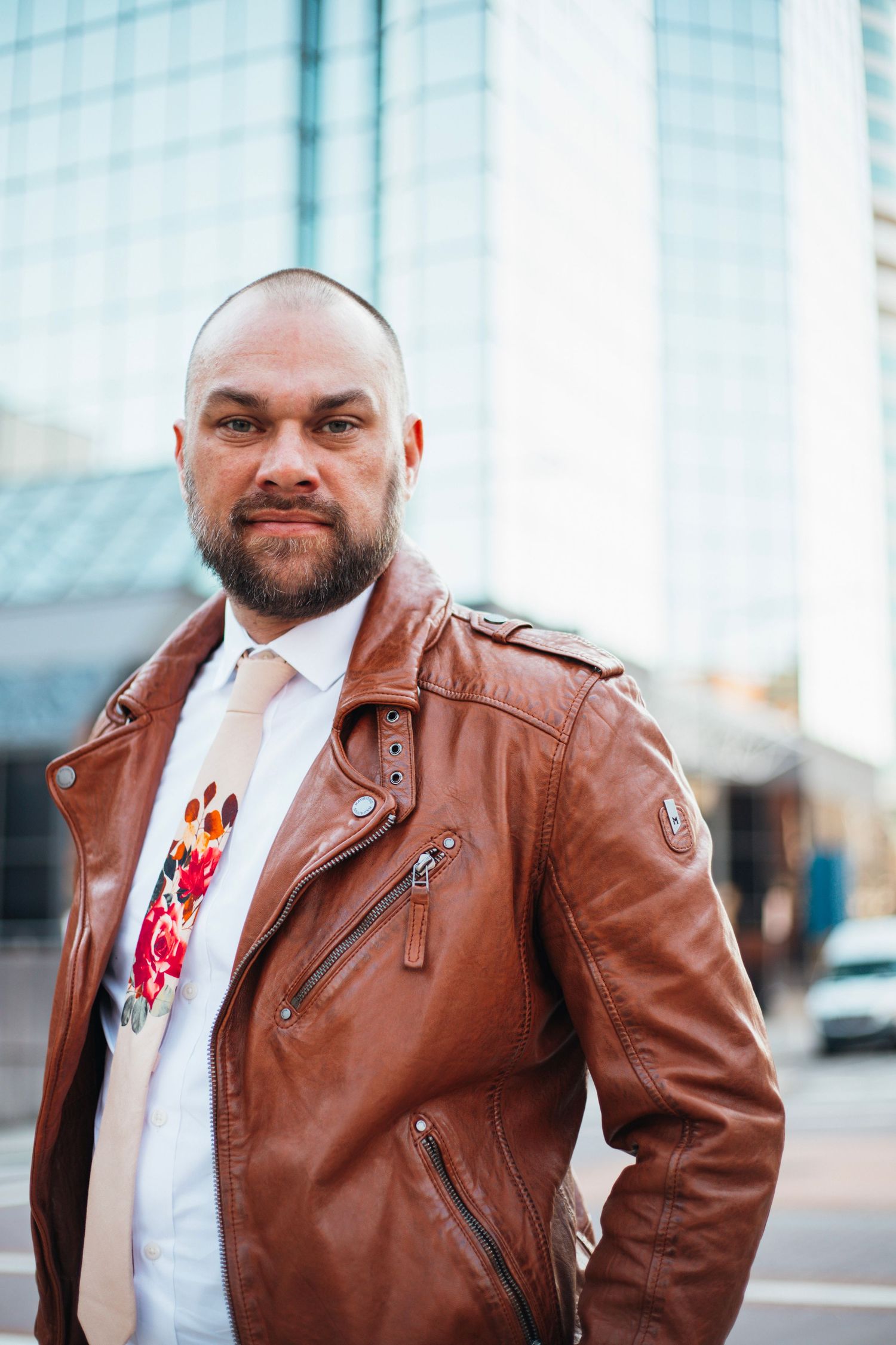 A person in a brown leather motorcycle jacket poses confidently in front of an urban glass building.