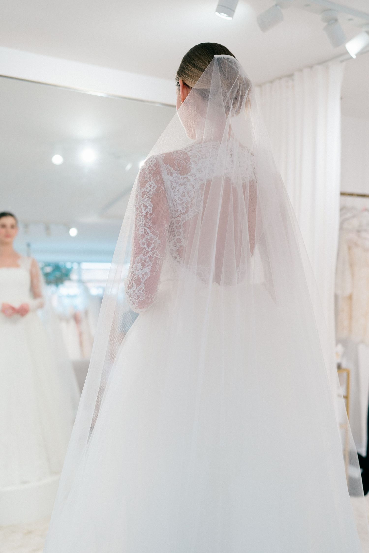 A bride in a lace-detailed wedding gown with flowing veil is photographed from behind in a bridal boutique.