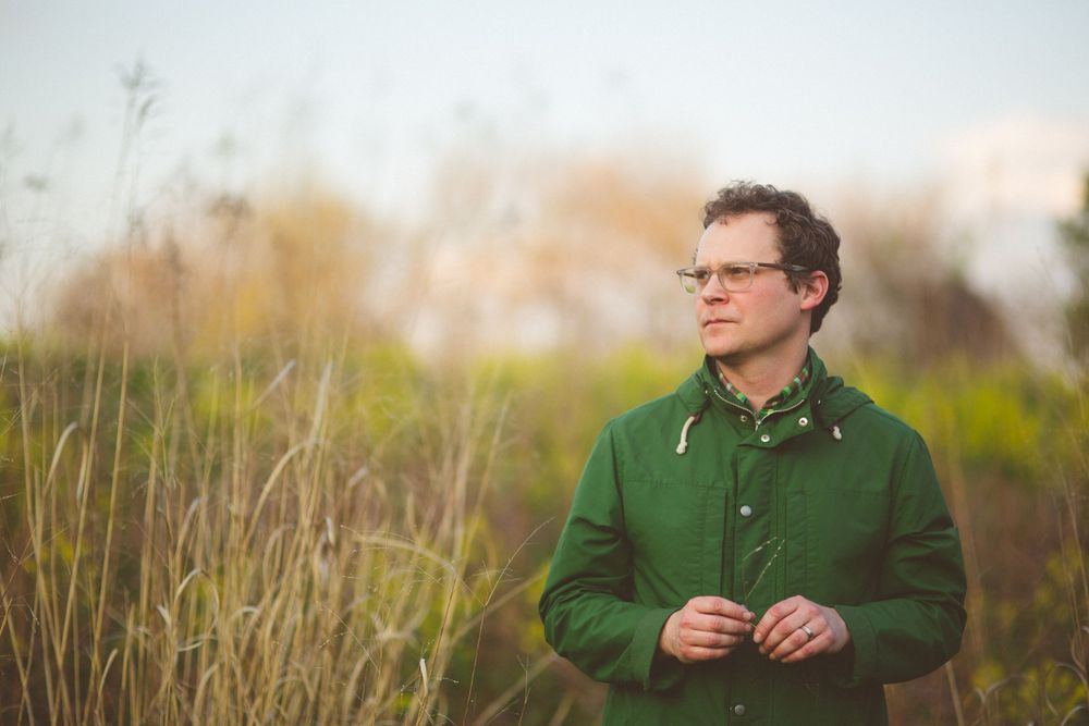 Man in green jacket standing thoughtfully in tall grass during golden hour.