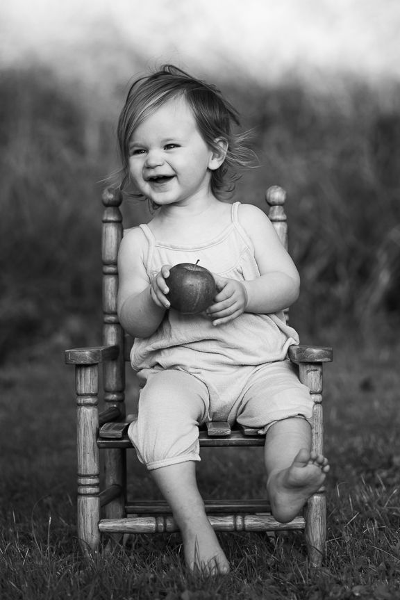 Black and white portrait of toddler laughing while holding apple in wooden chair.
