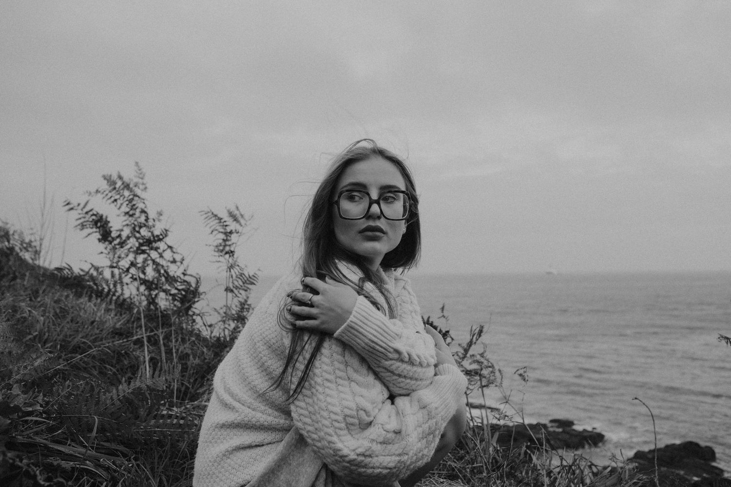 Person in glasses holding stuffed animal sits by coastal scenery in moody black and white photo.