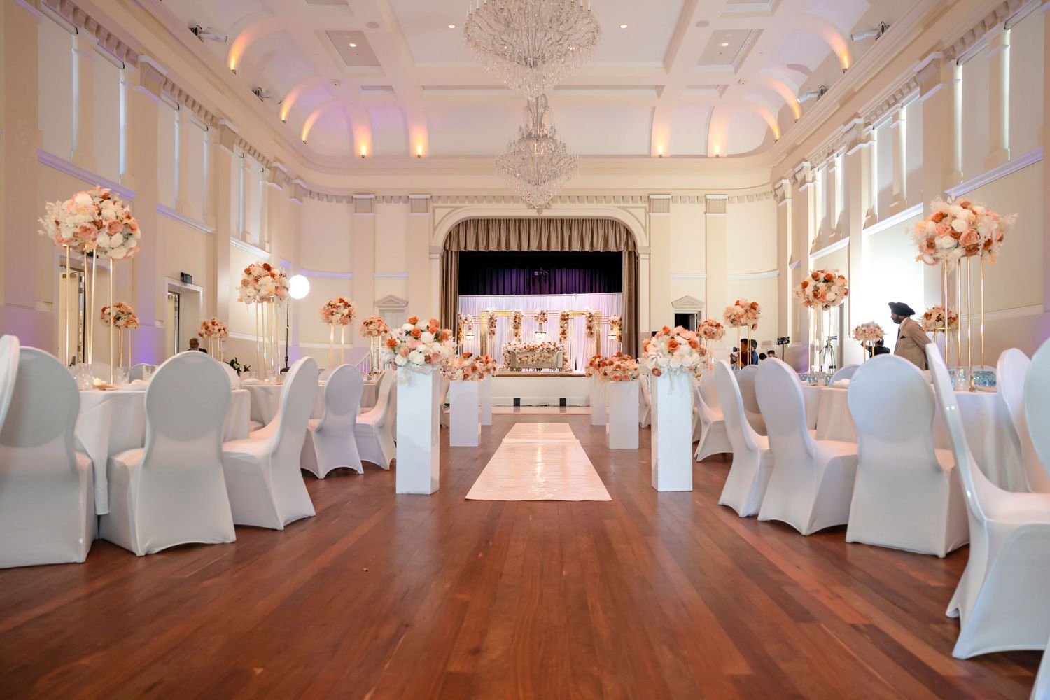 An elegant white wedding venue with chandelier lighting and chair covers leading to a decorated stage.