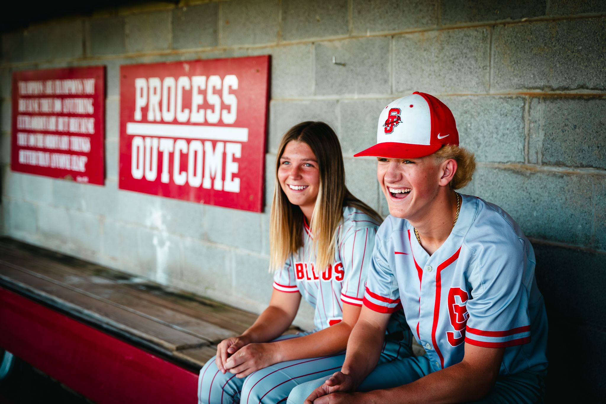 Two people sitting in dugout at baseball field with motivational signs on wall.