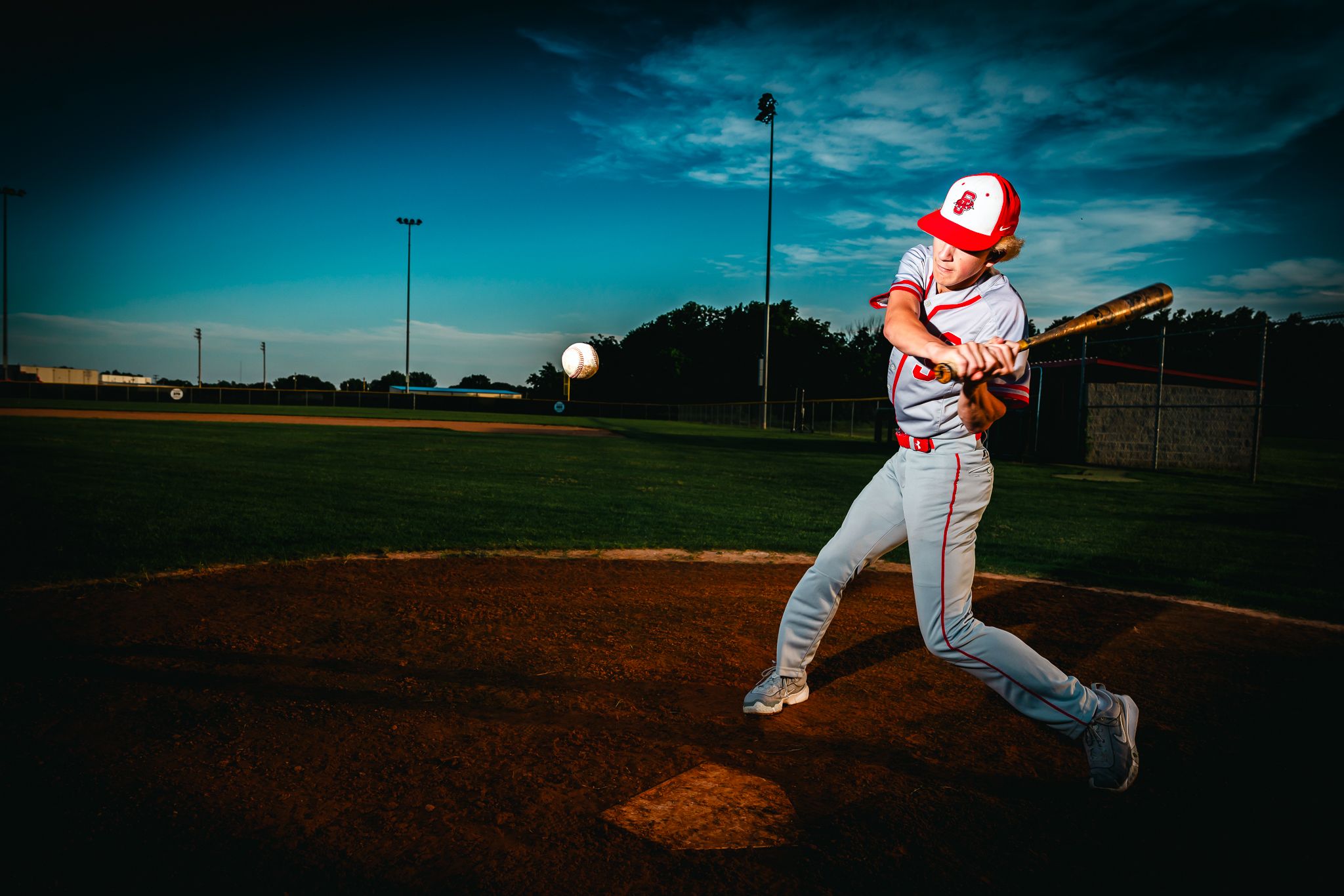 Baseball player in white and red uniform swings bat against dramatic blue sunset sky on baseball field.