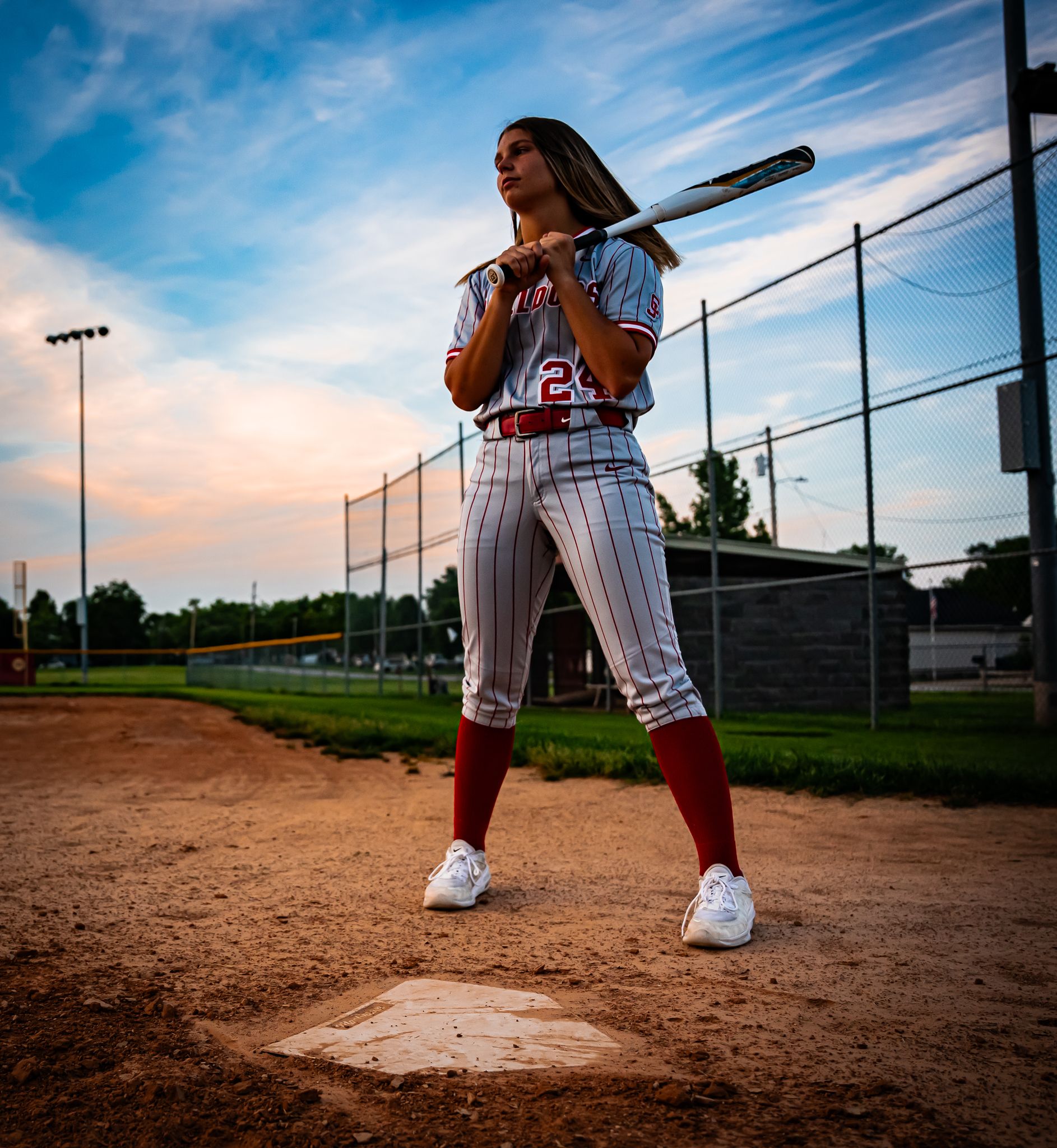 Baseball player in pinstriped uniform poses with bat on dirt field during sunset.