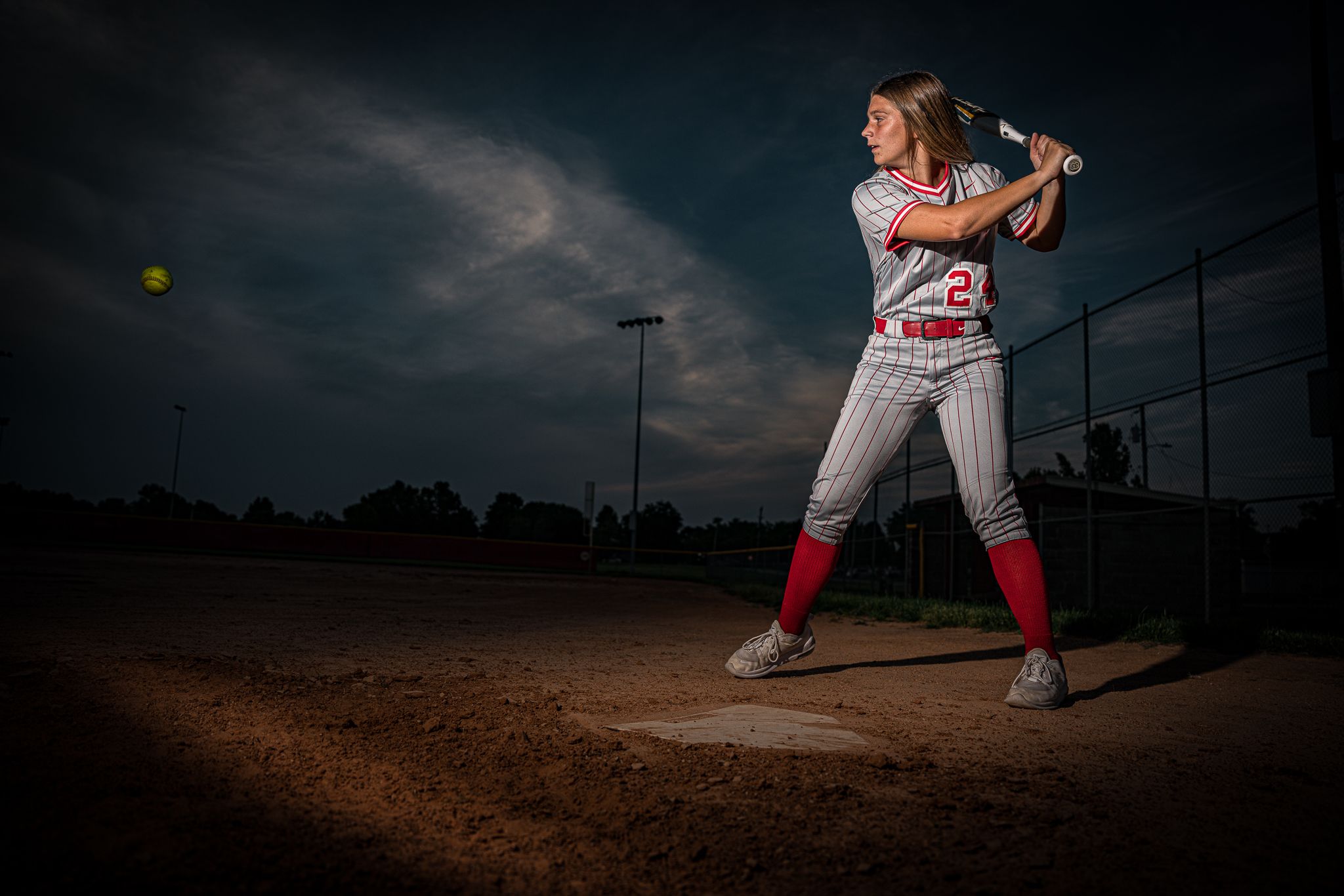 Softball player swings at yellow ball during nighttime practice on field.