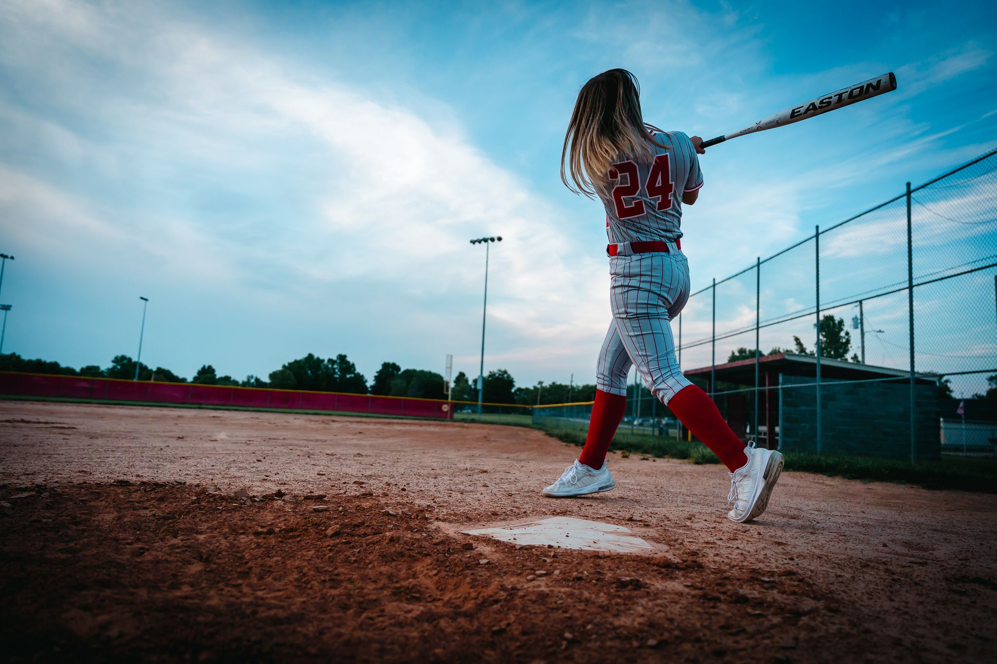 Baseball player in white uniform swings bat on dirt field against blue sky.