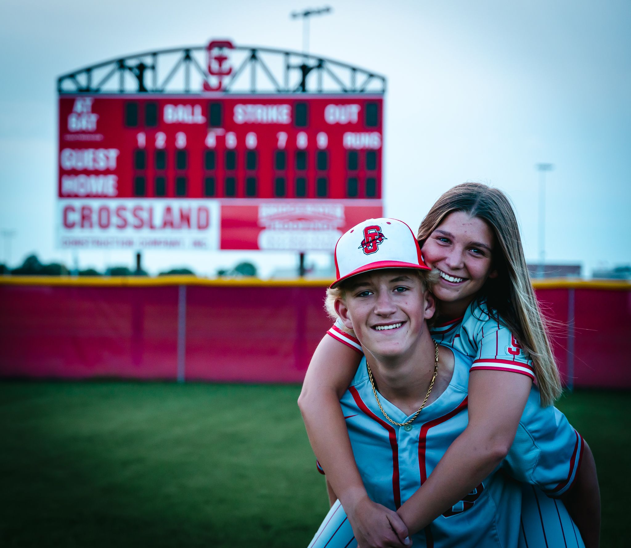 Two people embrace in front of red baseball scoreboard at dusk.
