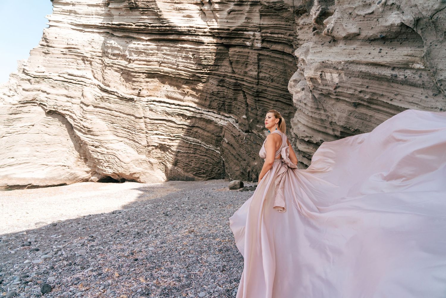 Flowing pink gown catches the wind dramatically against rocky coastal cliffs and natural stone formations.