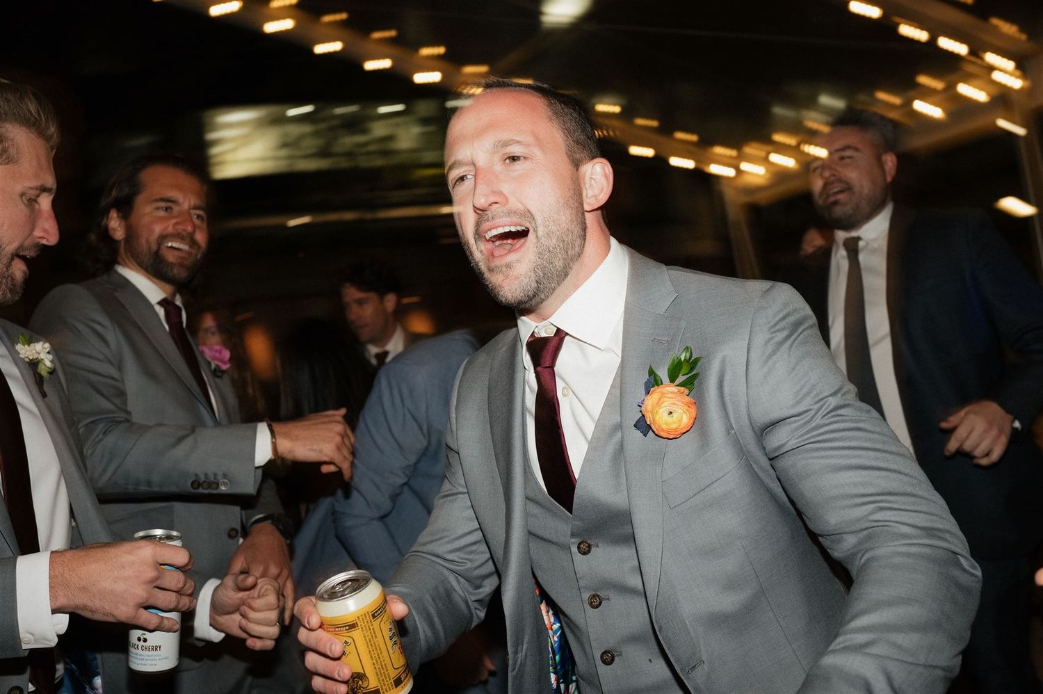 Wedding guest in grey three piece suit laughs while holding a drink at an evening reception celebration.