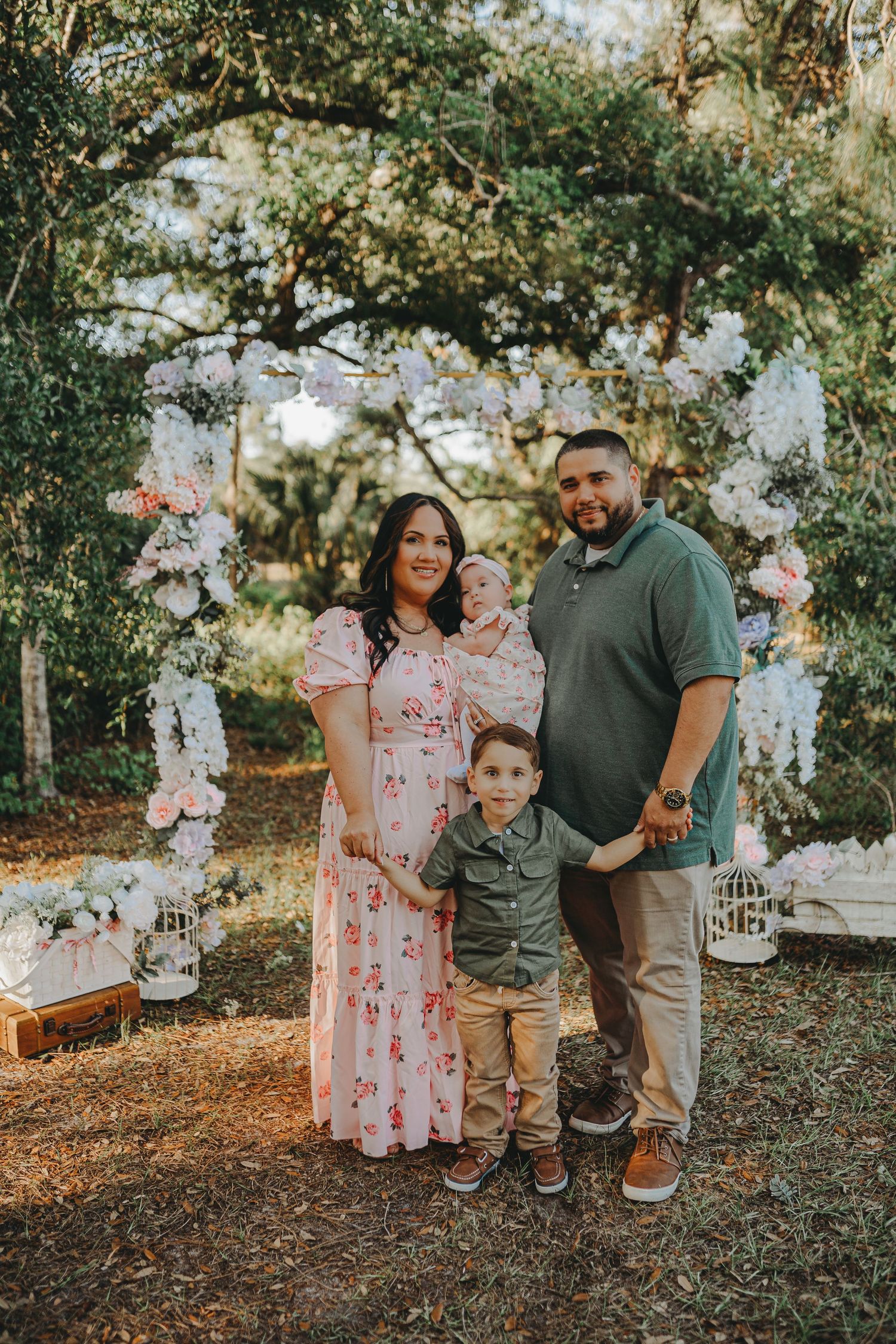 Family portrait under a white floral arch in a wooded outdoor setting.