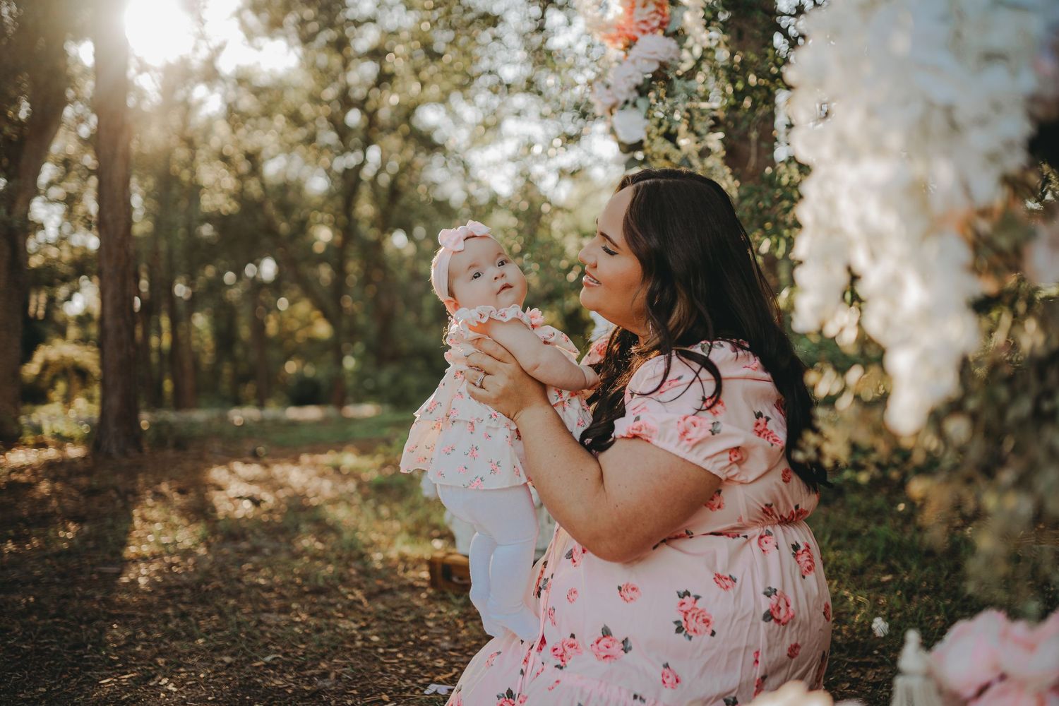 Sweet interaction between mother and child under floral arch with sunlight streaming through trees.