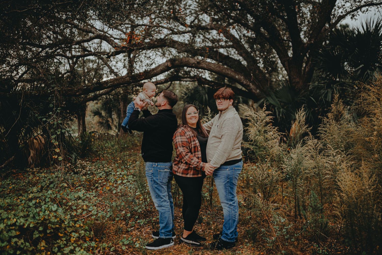 Casual outdoor family photoshoot with autumn foliage and arching tree branches in the background.