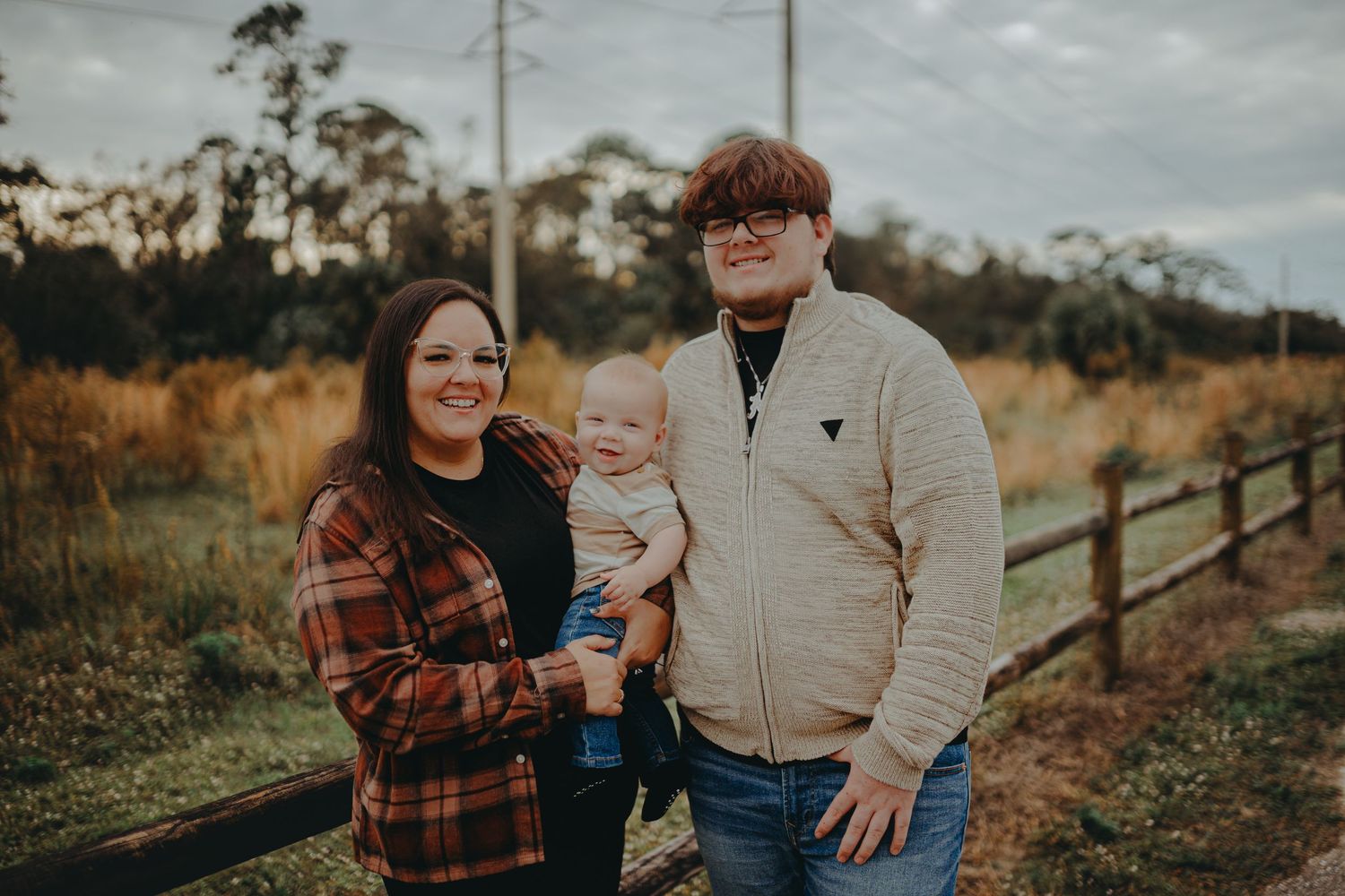 Rustic family portrait by wooden fence with cloudy sky and tall trees in background.