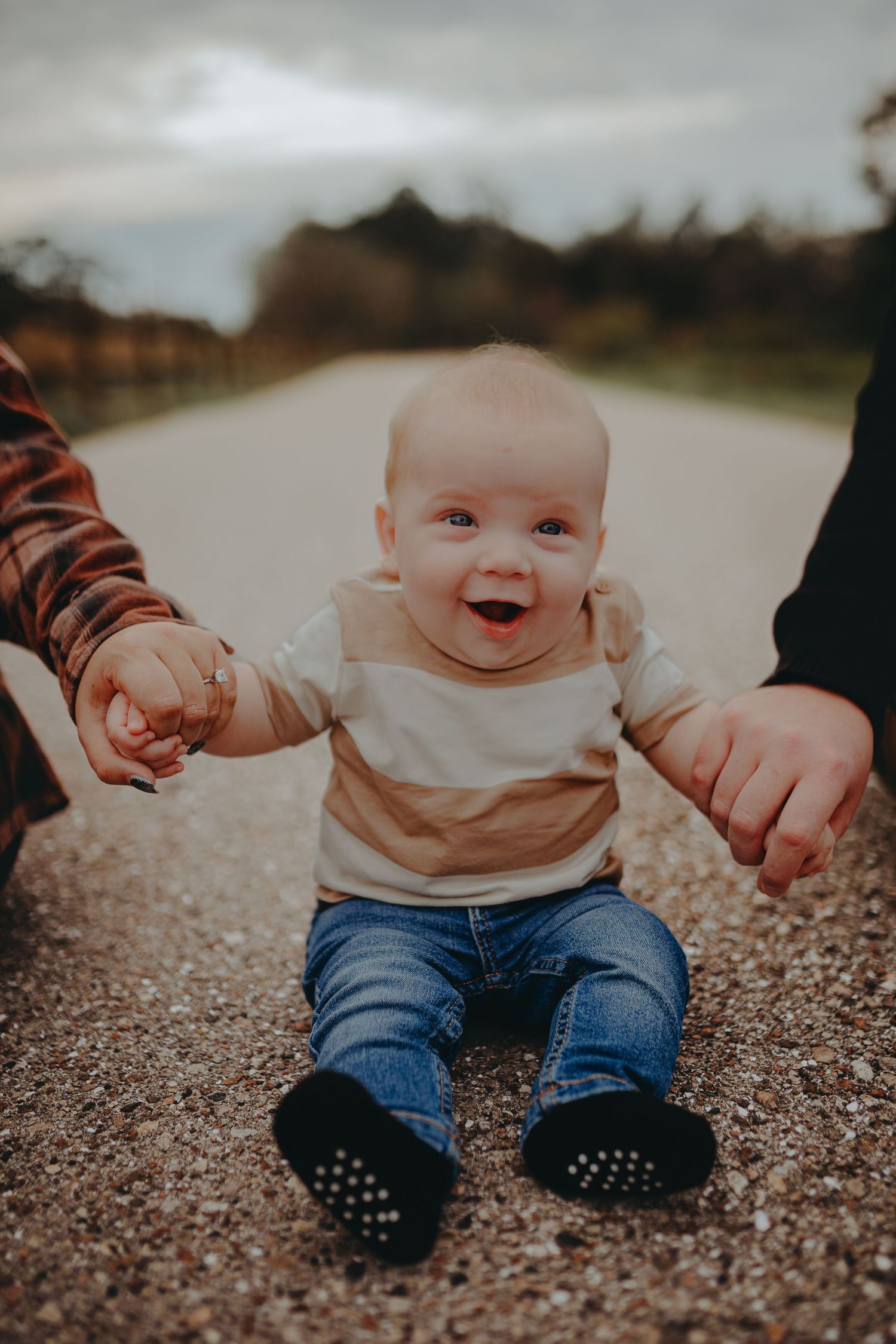 Happy baby sitting on gravel path wearing jeans and being held by parents' hands.