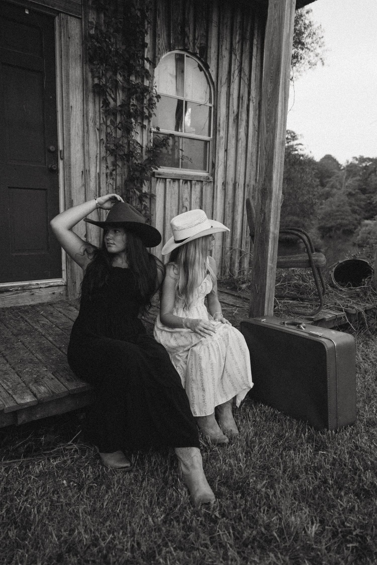 Two women in dresses and sun hats sit on wooden porch with vintage suitcase.