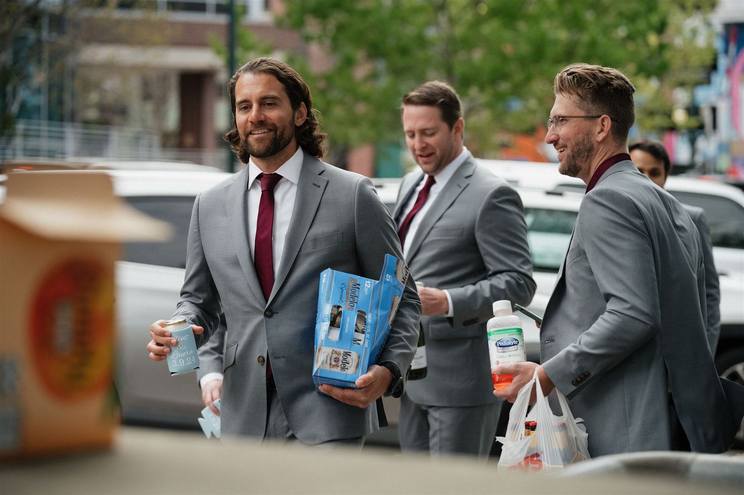 Three groomsmen in gray suits walking together before a wedding ceremony, with one carrying a bottle.