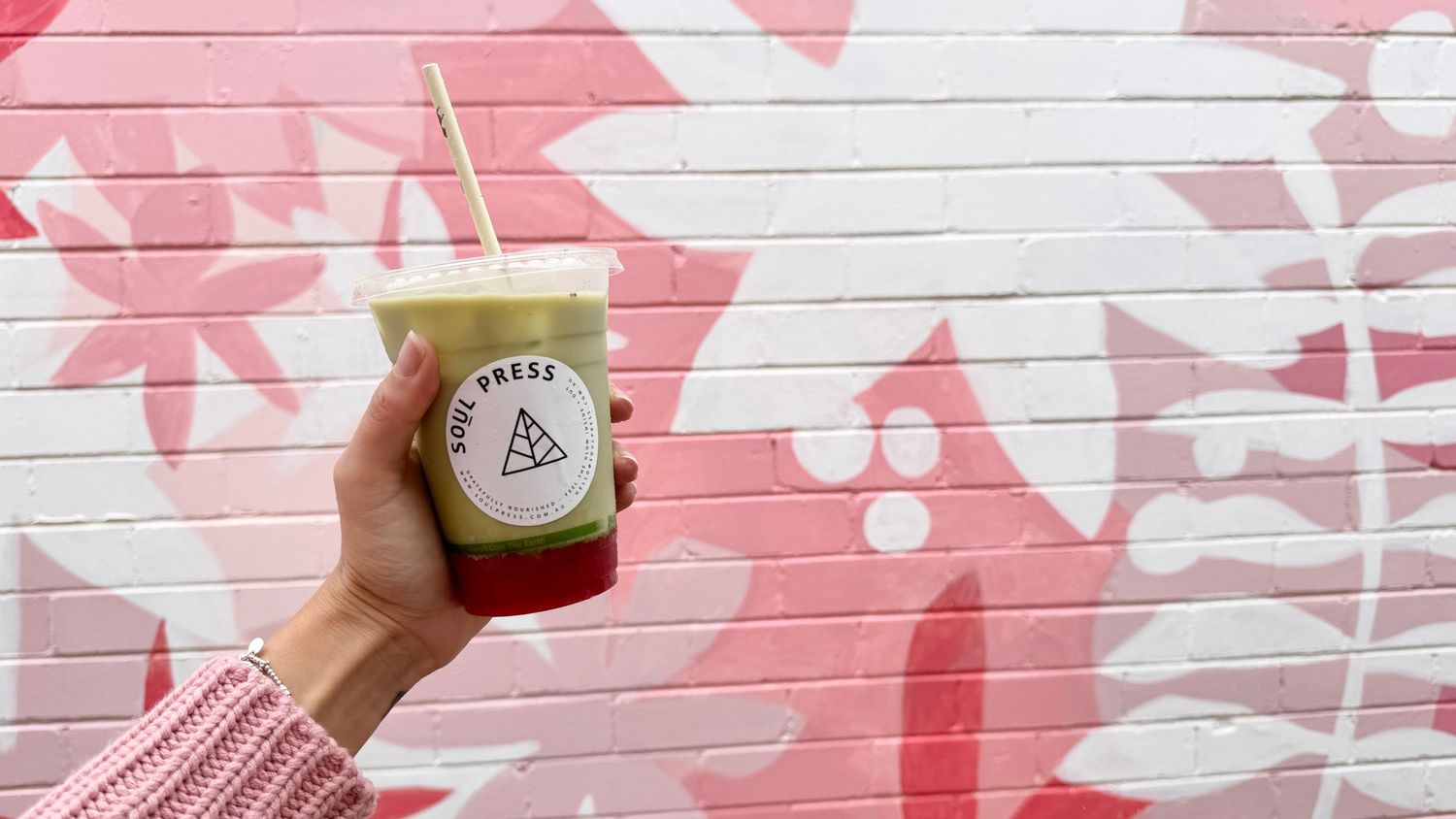 Hand holding Soul Press branded beverage against pink floral wall mural.