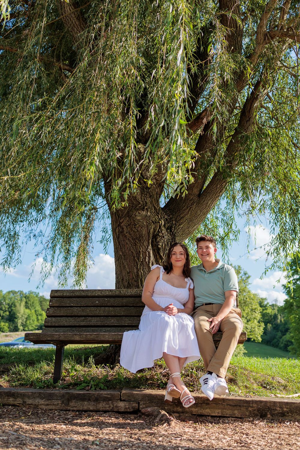 Couple relaxes on wooden bench under drooping willow tree branches.