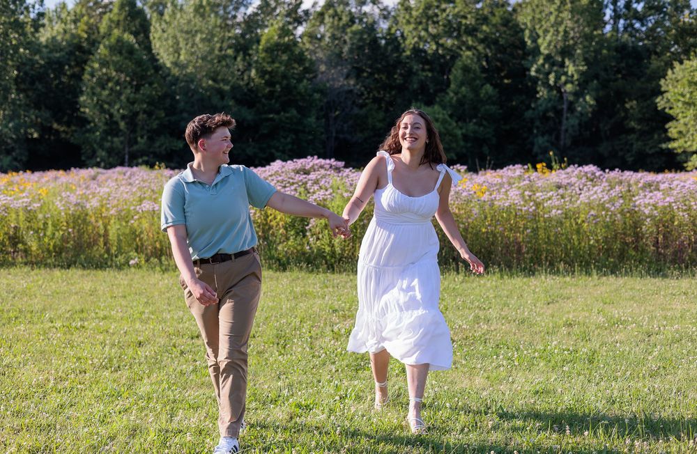 Couple runs playfully through sunny meadow with white dress flowing in the breeze.