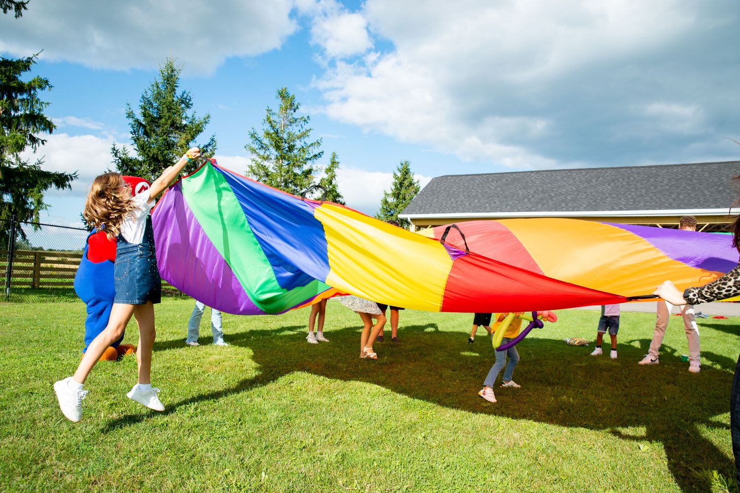 Group playing with colorful parachute on sunny day with green lawn and white fence.