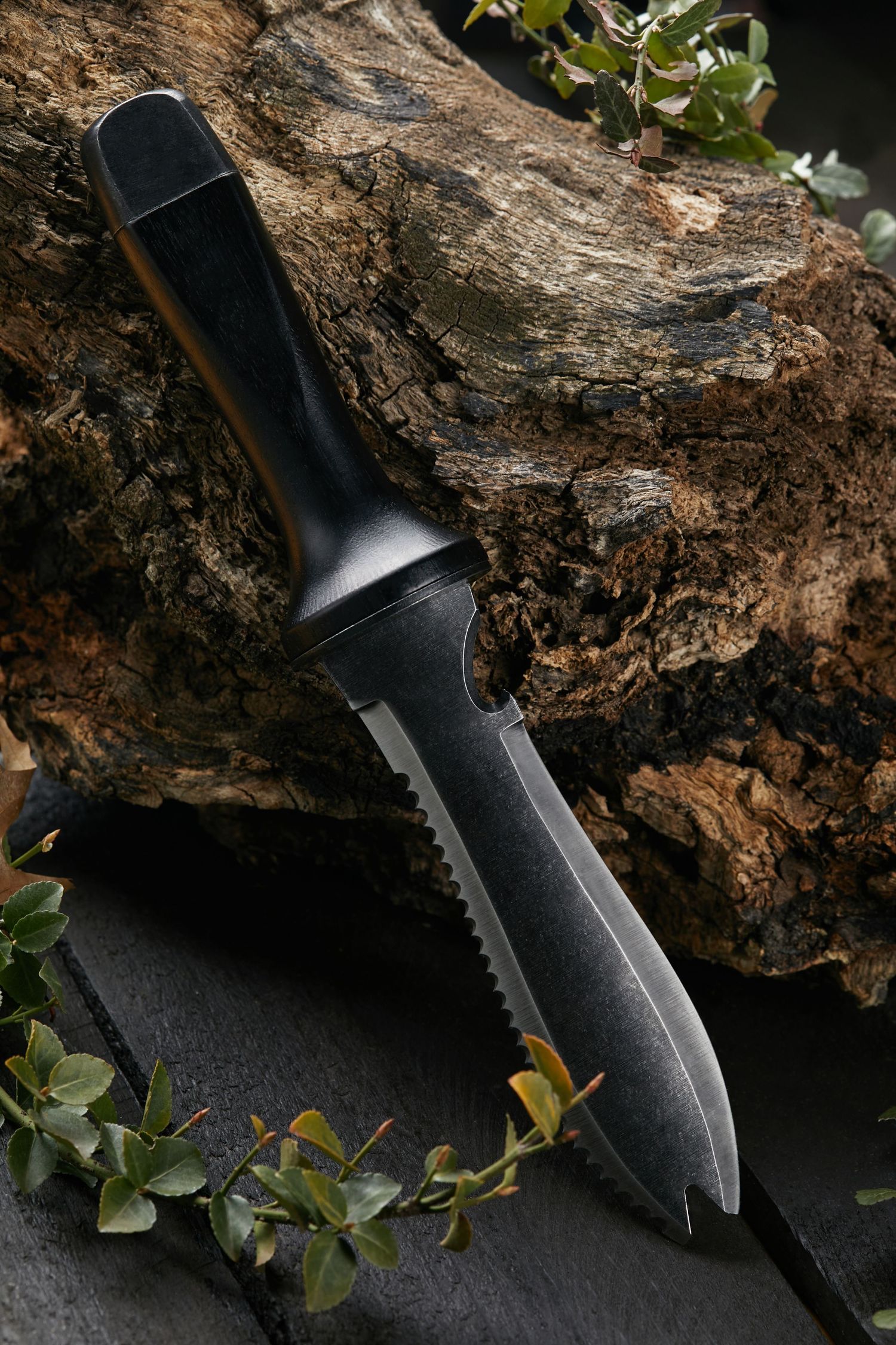 Black tactical knife photographed against textured bark with green foliage.Producing high-quality visuals for product advertisements