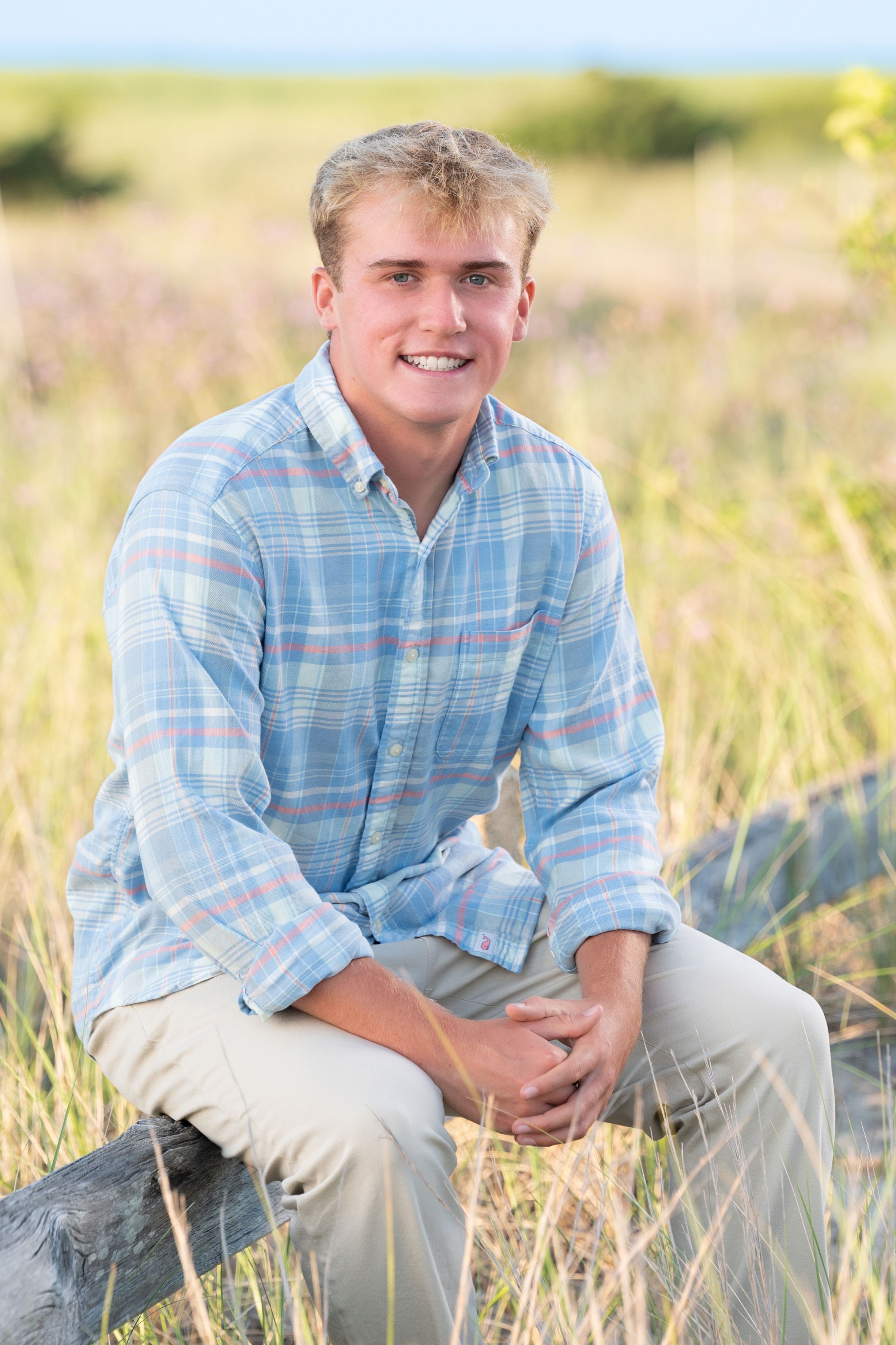 A person wearing a light blue plaid shirt sits in a sunlit grassy field during golden hour.